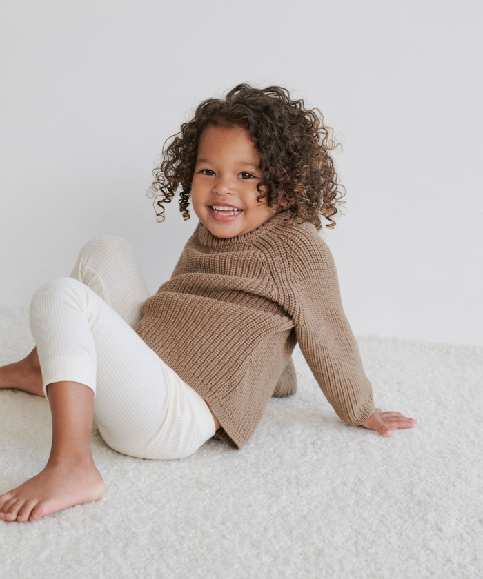 A young child with curly hair sits on a white carpet, smiling, wearing a cozy brown Kids Cotton Fisherman sweater and white ribbed leggings.