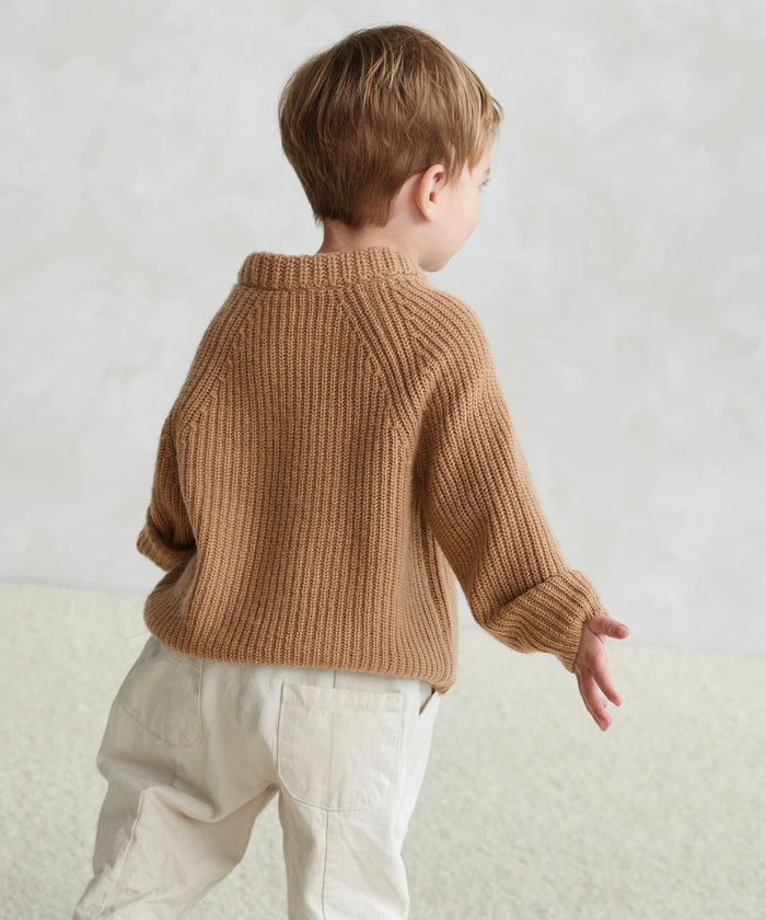 A young child with short light brown hair, seen from behind, wears the Kids Fisherman Sweater and off-white pants while standing on a textured floor against a pale, softly patterned background.