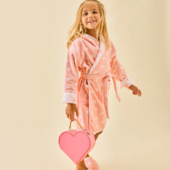 Smiling girl holding a heart-shaped bag and wearing a fluffy coral pink bathrobe with white hearts and matching slippers.