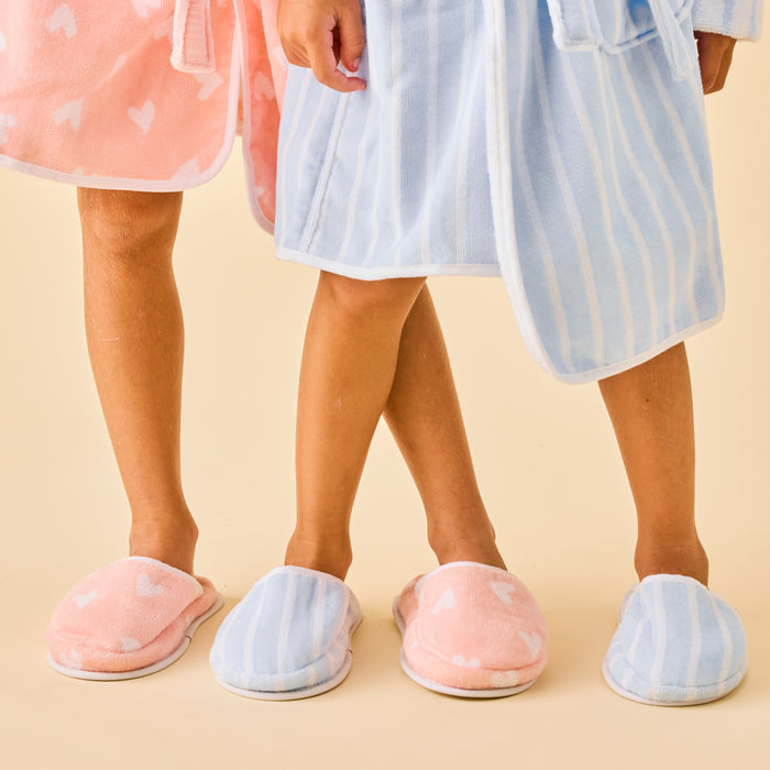 Two kids wearing soft terry slippers and matching bathrobes in coral hearts and blue stripes.