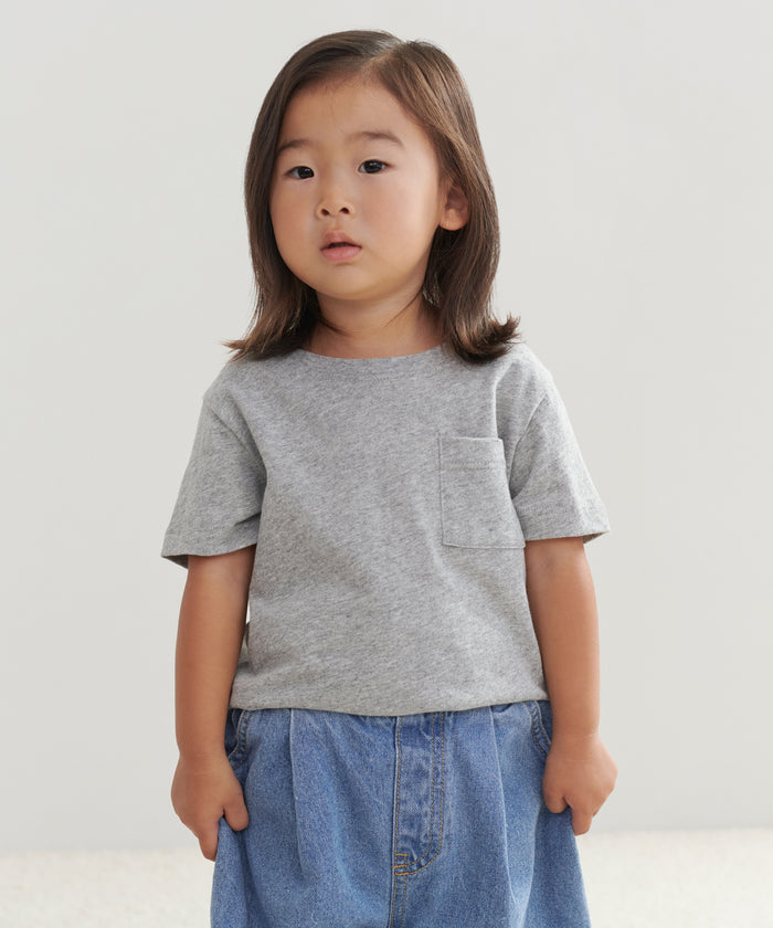 A young child with straight brown hair wears a gray Kids Vintage Pocket Tee and a blue denim skirt, standing against a plain light background and looking slightly to the side.