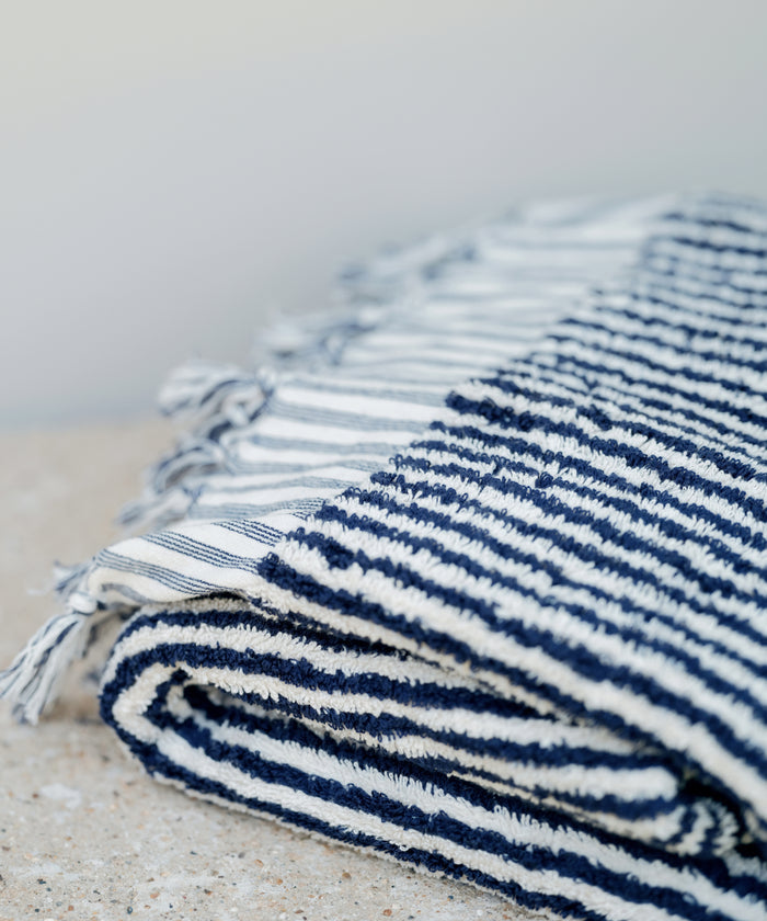 A Laguna Beach Towel, made from Turkish cotton with navy blue and white stripes plus tasseled edges, is folded and placed on a light-colored surface.