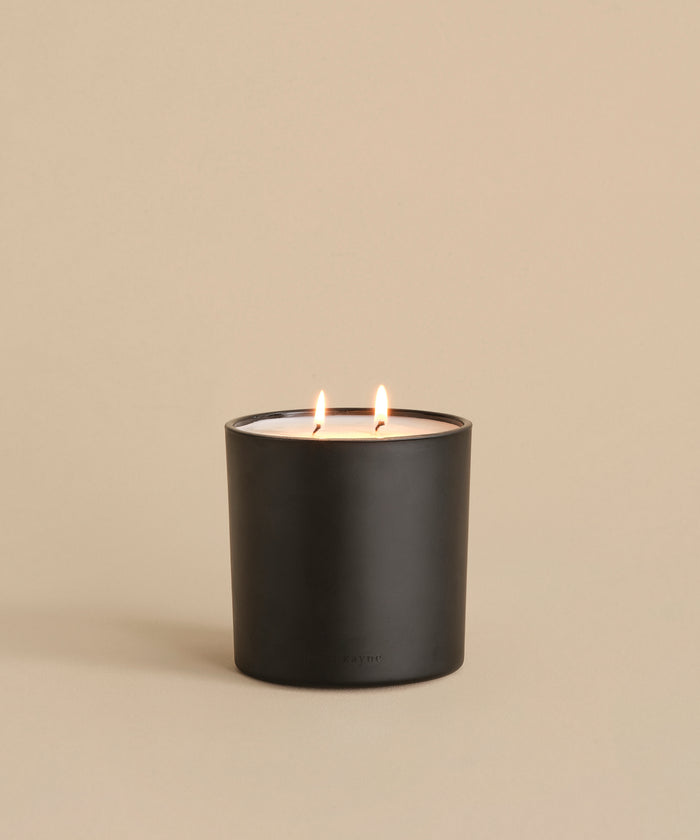 The Large Aspen Glass Candle, with two lit wicks, sits on a neutral beige surface and casts a soft glow against a plain beige background.