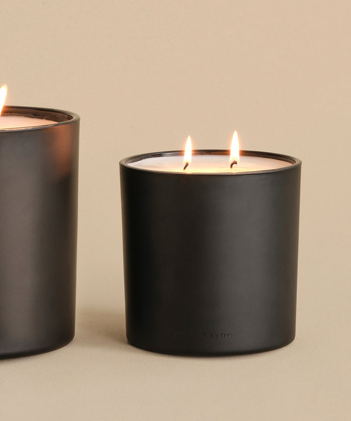 A large black Large Aspen Glass Candle with two lit wicks rests on a neutral surface, while another similar candle is partially visible on the left. The background is plain beige.