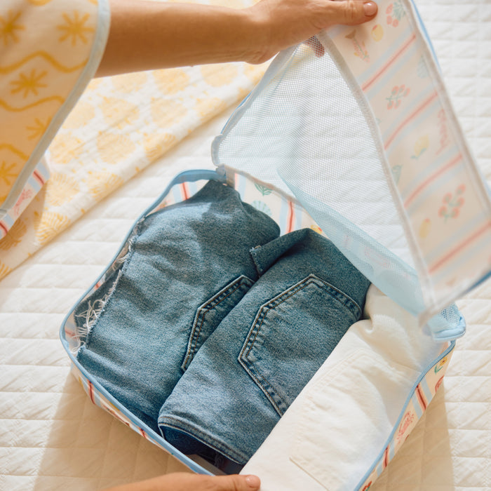 A person’s hands are closing the mesh lid of a Large Packing Cube in the Dolce Vita print, which features a light blue background with a colorful pattern of lemons and tomatoes. Inside the open cube, folded denim jeans and white fabric are neatly packed, resting on a white quilted surface.