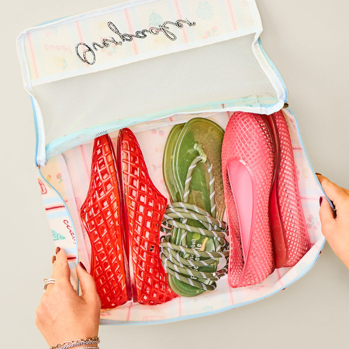 Hands holding a Dolce Vita packing cube filled with colorful jelly sandals and a pair of rope sandals on white surface.