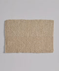 The Laurel Bath Mat, a rectangular, woven beige mat made from 100% fique, lies flat on a light gray surface. Its coarse texture with visible fibers and a faint horizontal line gives it an ivory, natural look.