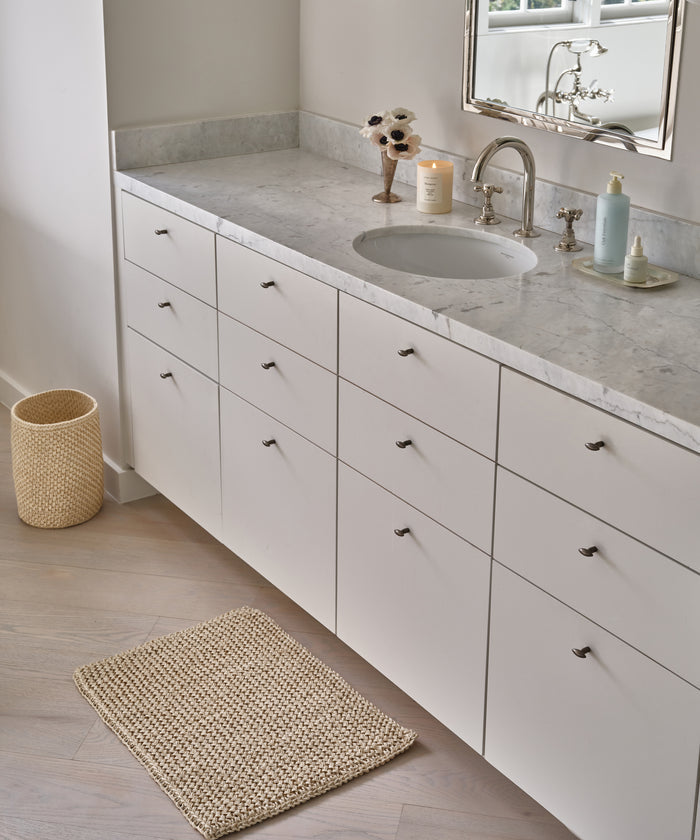 Modern bathroom vanity with white drawers and marble countertop, featuring a round sink, silver faucet, soap dispenser, candle, floral decor, woven trash bin, and the Laurel Bath Mat made from 100% fique on a light wood floor.