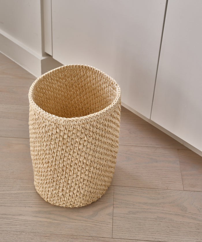The Laurel Waste Basket in beige, handwoven and natural, rests on a light wood floor beside white cabinets, bringing organic charm to a modern minimalist space.