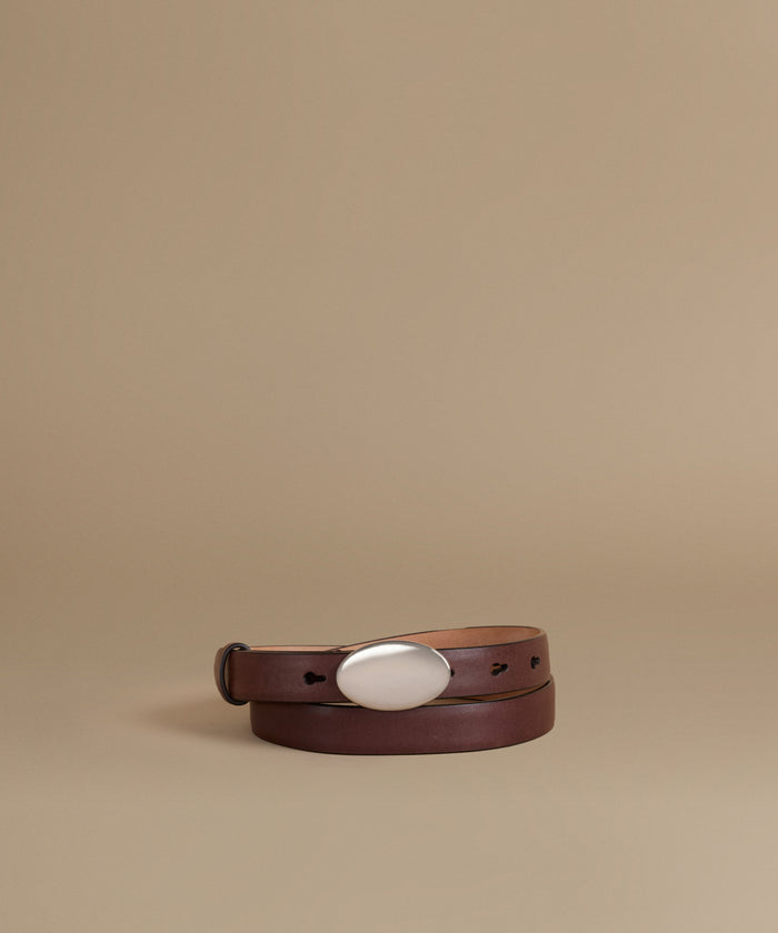 A dark brown leather belt with a large, polished silver oval buckle, neatly coiled on a warm beige studio background.