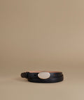 A black leather belt with a large oval white mother-of-pearl style buckle, coiled on a warm beige studio background.
