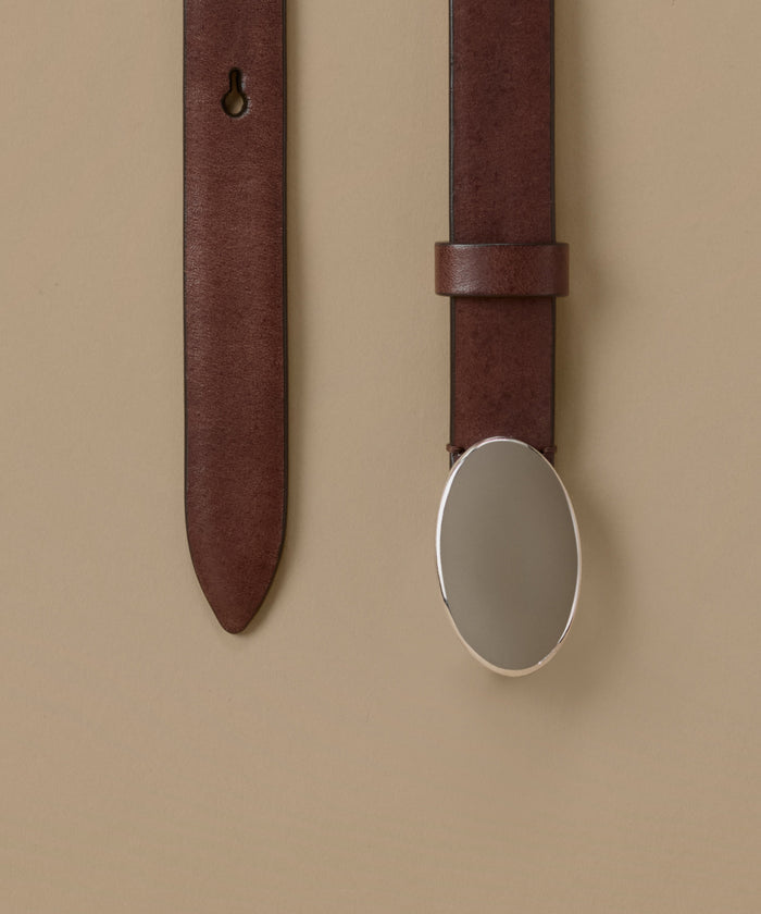 A close-up of a dark brown leather belt with a large, polished silver oval buckle, shown against a solid beige background.