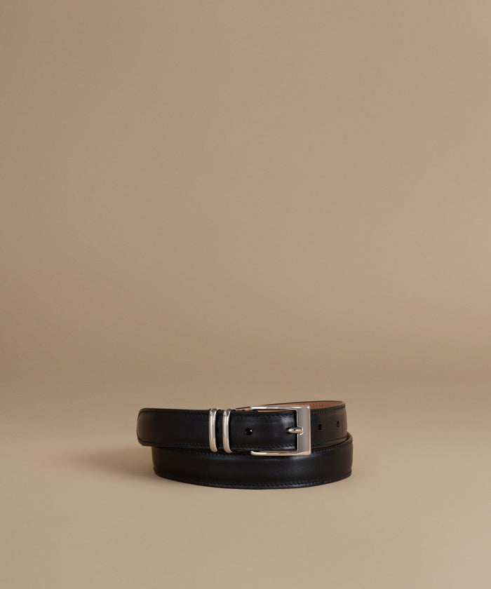 A black leather belt with a silver-tone buckle, neatly coiled on a warm beige studio background.