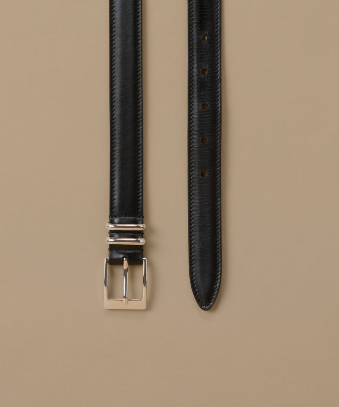 A black textured leather belt with a polished silver-tone buckle, shown laid flat against a solid beige background.