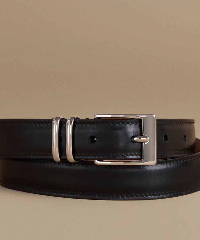 A black leather belt with a polished silver-tone buckle, displayed in a coiled arrangement on a solid beige background.