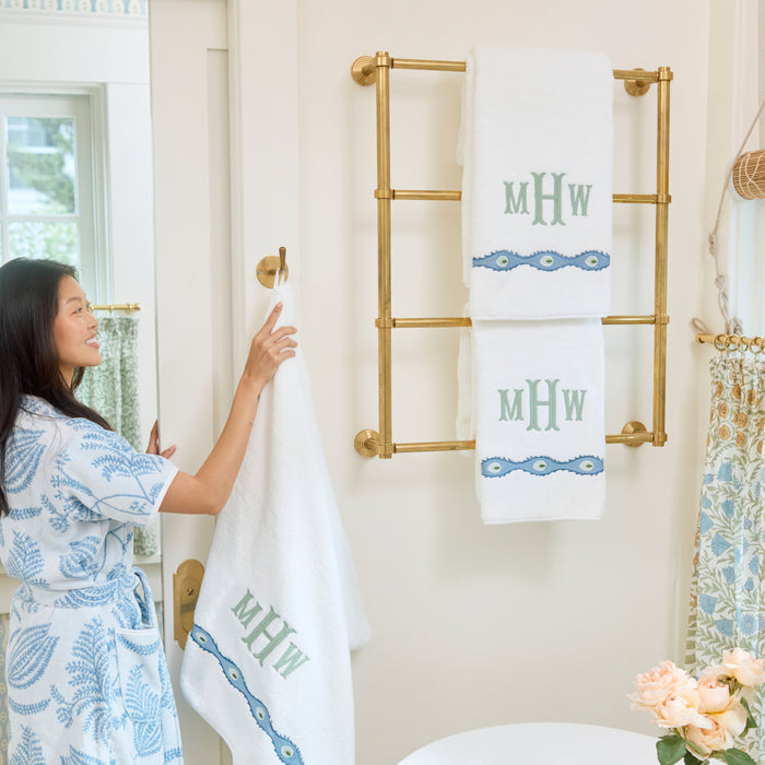 Woman in a blue patterned robe hangs a white bath towel with embroidered motif and a green MHW monogram on a brass rack.