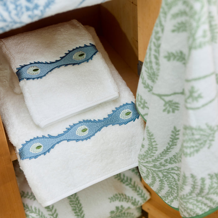 Folded white bath towels with blue wavy embroidery on a wooden shelf next to a green leafy towel.