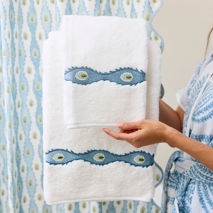 Person in a blue patterned robe holding folded white bath towels with blue and green embroidery.