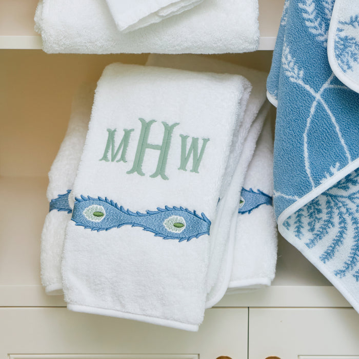 White Weezie hand towel with green MHW monogram and blue Lee Jofa trim on a white shelf.