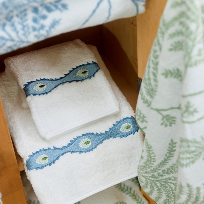 Stacked white Weezie hand towels with blue Lee Jofa embroidery inside a light wooden cabinet.