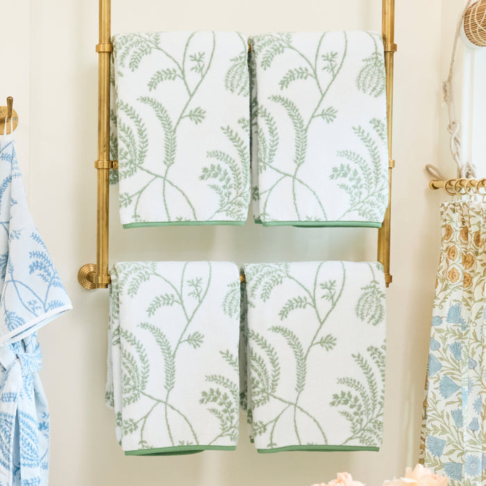 Four folded white bath towels featuring a sage green Palmero pattern on a gold towel rack.