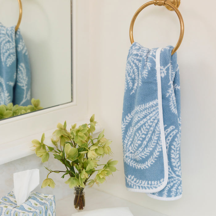 Patterned light blue hand towel hanging on a round brass ring next to a vase of green flowers.
