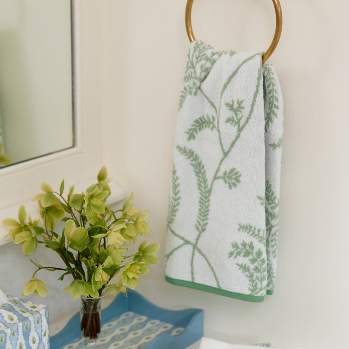 Lee Jofa Palmero Sage hand towel hanging on a brass ring next to a vase of green flowers.