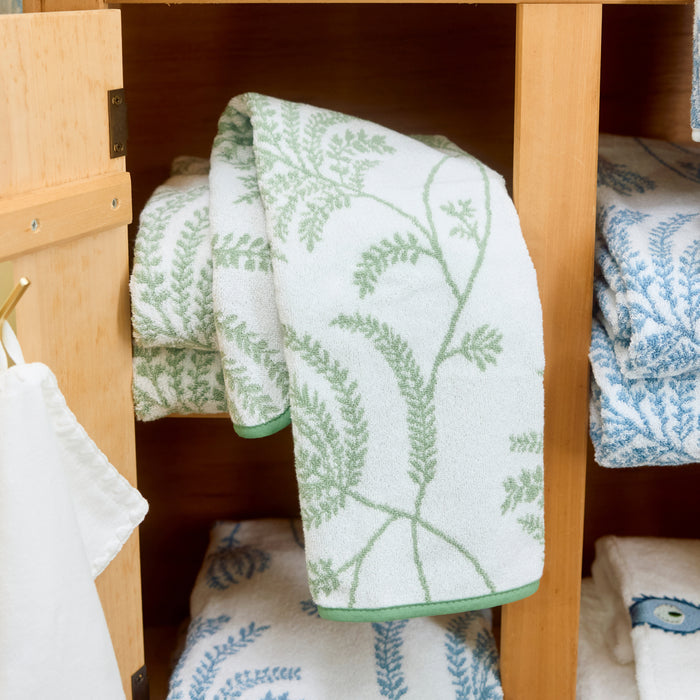 Folded white hand towel with sage green Palmero botanical pattern draped over a wooden shelf.