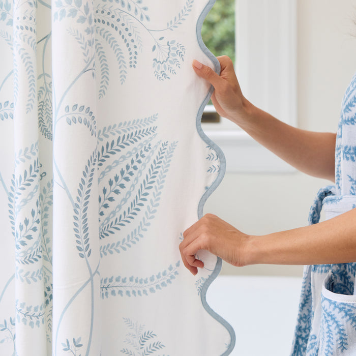 Hands adjusting the white Lee Jofa Palmero Sky shower curtain with a blue scalloped edge.
