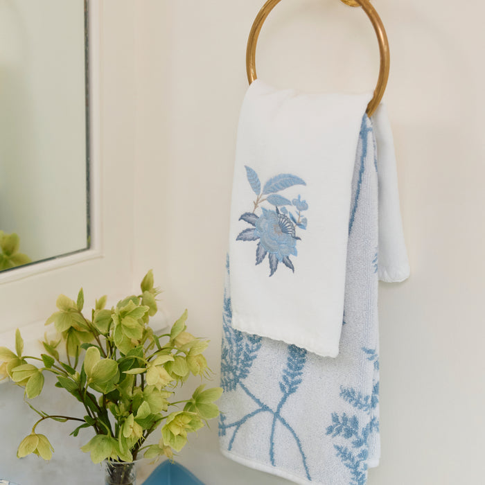 White bath towel with blue floral embroidery hanging on a brass ring above green flowers.