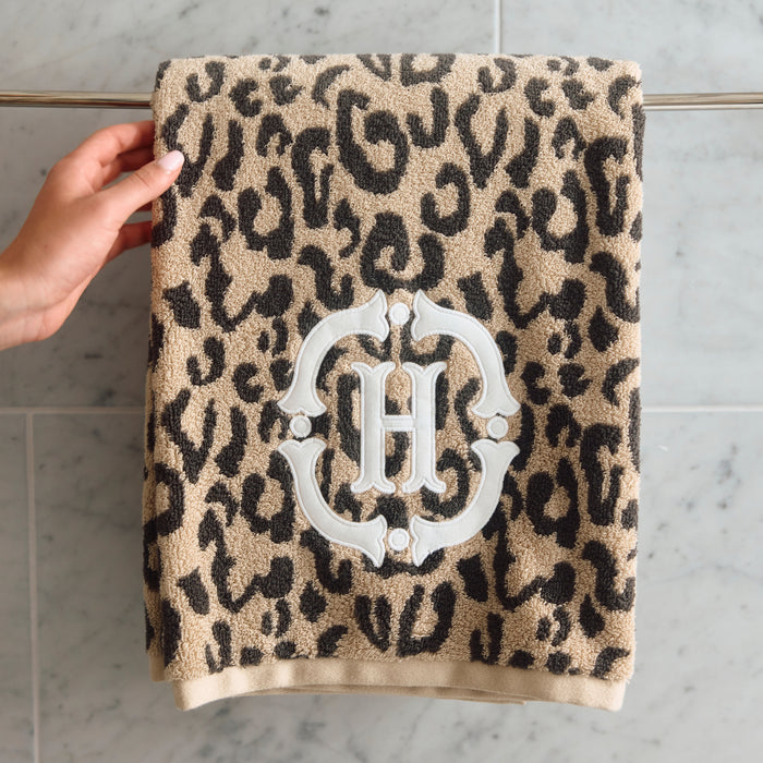 A beige and black leopard print bath towel with a white monogrammed 'H' crest, hanging from a metal towel bar on a gray marble tiled wall. A hand is visible holding the left side of the towel.