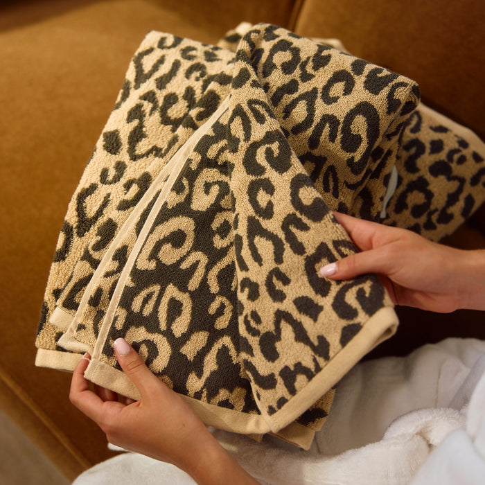 A close-up of a leopard print bath towel being held by hands, showing its soft terry cloth texture and beige border, set against a warm brown background.