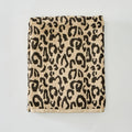 A folded cotton towel with a classic leopard print pattern in black on a warm beige background, featuring a solid beige border along the bottom edge, photographed flat against a light gray background.