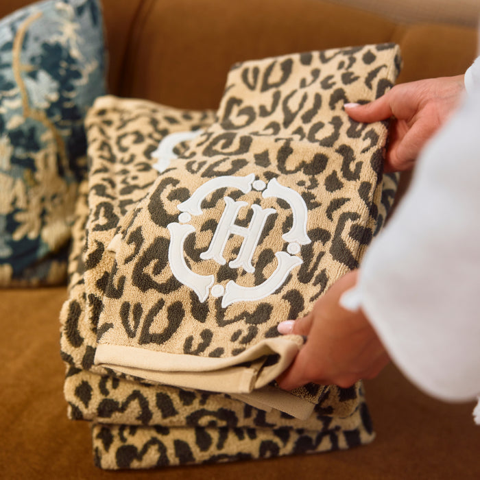 A stack of leopard-print bath towels being held by hands, featuring a white monogram 'HJ' on the top towel, set against a brown leather sofa background.