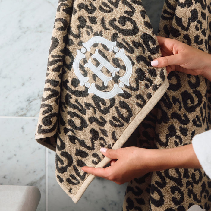 A close-up of a leopard-print bath towel being held by hands, showing a beige and black animal pattern with a white embroidered monogram logo.