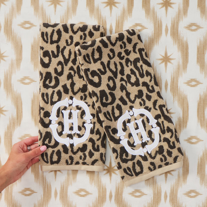 A pair of leopard-print bath towels laid flat on a patterned fabric background, with a hand holding one corner of the left towel. Each towel has a large white decorative monogram letter H near the bottom.