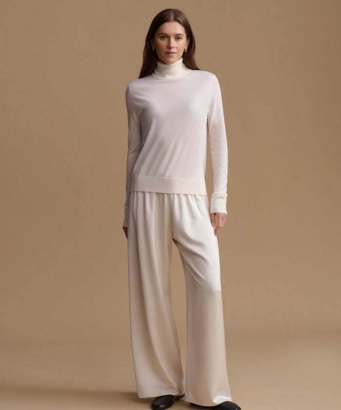 A woman modeling a cream-colored knit turtleneck sweater and matching wide-leg trousers against a solid beige studio background.