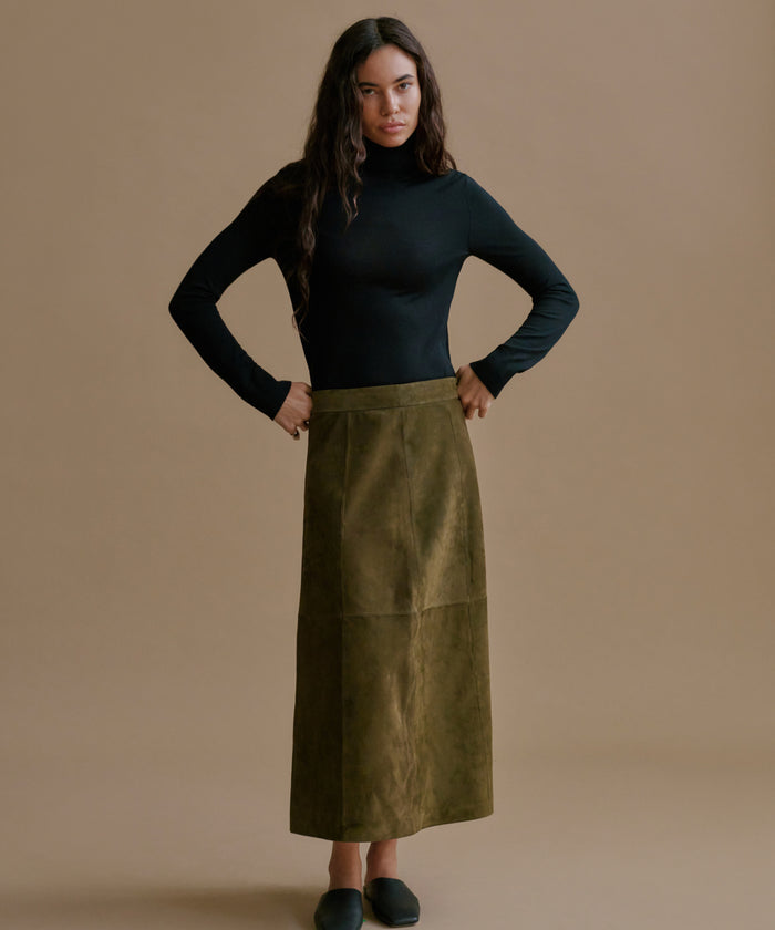 A woman stands against a plain beige studio background wearing a fitted black long-sleeve turtleneck top and an olive green midi skirt. She is posed with hands on hips, wearing dark slip-on shoes.