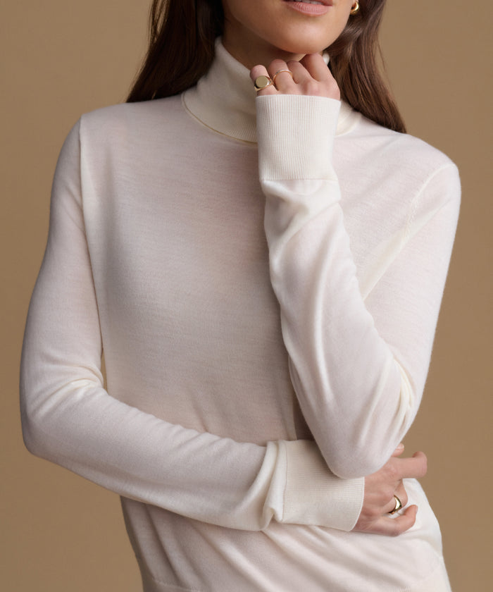A close-up of a woman wearing a cream-colored, long-sleeve mock neck knit sweater against a solid warm beige background.