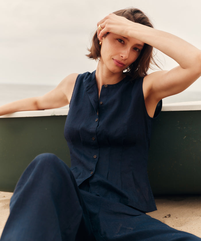 Woman wearing a navy blue sleeveless top with blue pants sitting on a ledge.