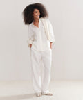 A woman with voluminous curly hair smiles in a loose white button-up, Linen Logan Pant in ivory, open-toe sandals, and a draped sweater—an effortlessly stylish everyday look.