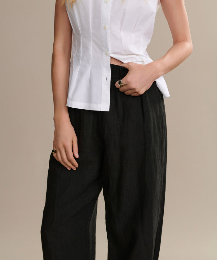 Wearing the Linen Logan Pant and a sleeveless white button-up top, a person poses with one hand in their pocket against a beige background—showcasing an easy, versatile look for everyday wear.