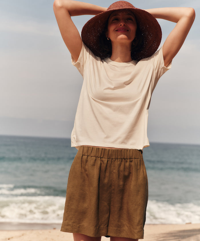 Woman wearing a wide-brimmed hat and beige top with brown shorts on a beach.