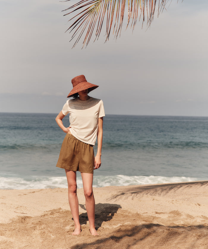 Person standing on a beach wearing a wide-brimmed hat and shorts, with palm leaves in the background.