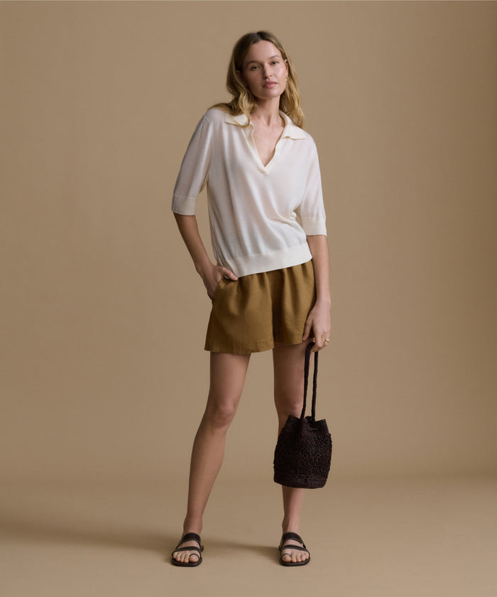 Woman wearing a white shirt and brown shorts, holding a brown bag against a beige background