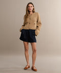 A woman poses against a beige background wearing the Logan Short in black with a tan jacket and brown flats. Her long hair frames her face as she stands with hands in her pockets, showcasing the shorts’ lightweight comfort.