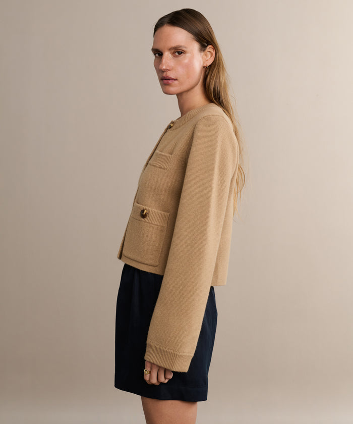 A person with long hair stands sideways in a tan, long-sleeved, cropped jacket with gold buttons and black Logan Short against a plain beige background.