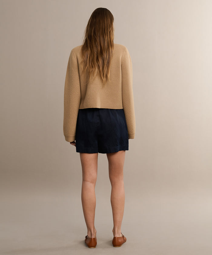 A woman with long hair faces away from the camera, wearing a tan long-sleeve top and the Logan Short, made from lightweight fabric with a subtle satin-like finish, against a plain light background.