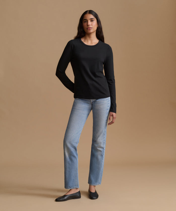 A woman modeling a black long-sleeve crewneck top with light-wash blue jeans and black flats, standing against a solid beige studio background.