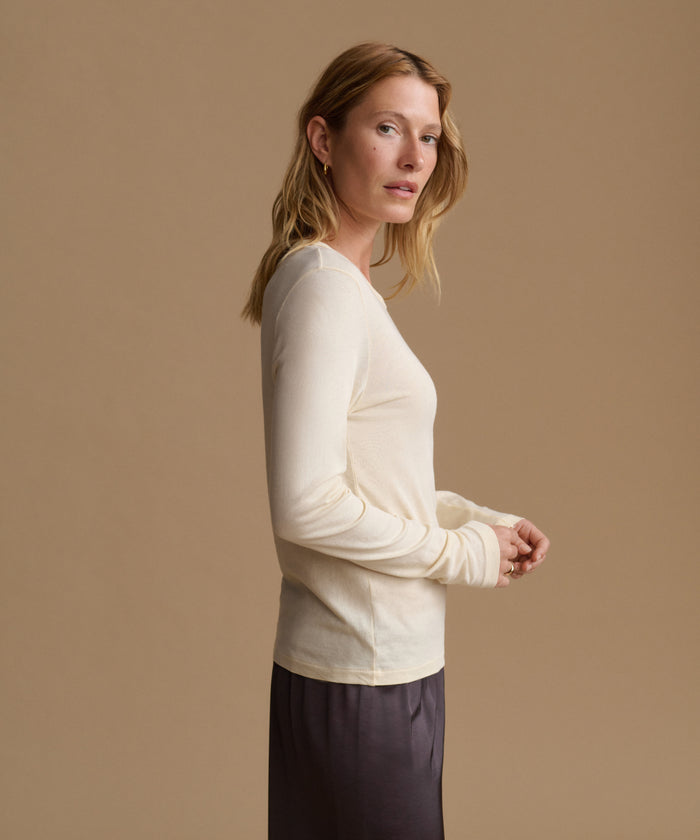 A woman in a side profile wearing a cream-colored long-sleeve knit top and a dark gray skirt, standing against a solid beige studio background.